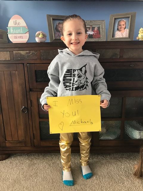 Michaela is smiling, wearing a grey sweatshirt with her school’s mascot that reads “Wildcat Pride” and holding a bright yellow sign that says “I miss you.  Love, Michaela”.