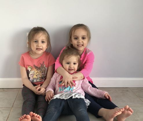 Sitting against a wall with their legs stretched front,  the three Lopatofsky sisters pose with smiles and laughter.  Michaela pictured on the left sits beside her younger sister Alexandra on the right, who wraps her arms around her baby sister Aurora sitting  front of her big sisters.